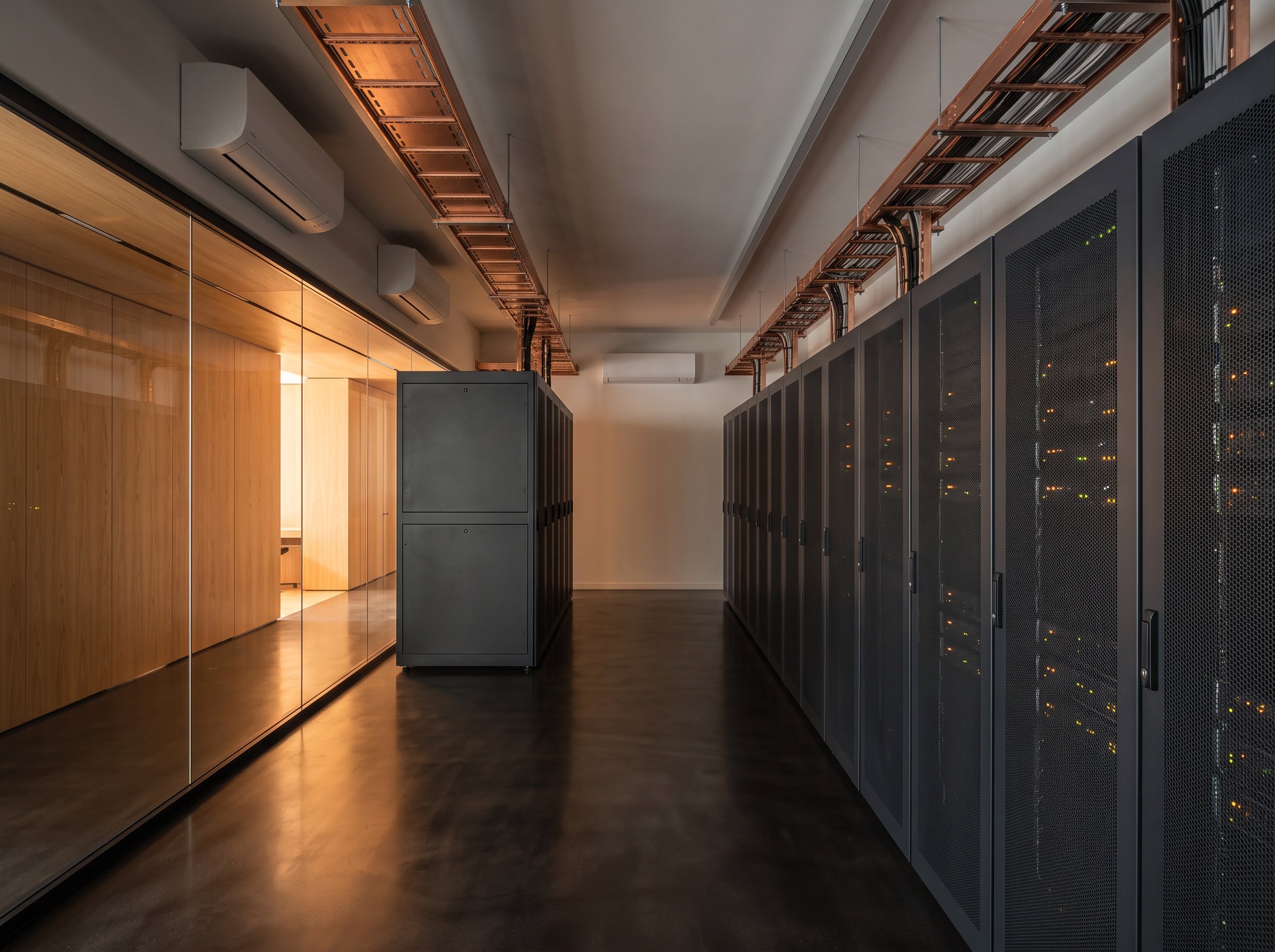 Zwei Klimageräte hoch an der Wand über einer Reihe dunkler Serverschränke, darüber Kabeltrassen, links die verglaste Wand zum Büro.